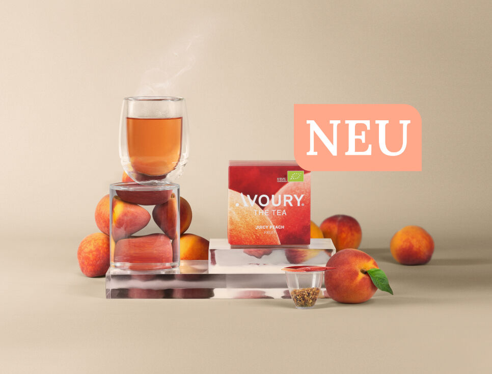 Eine Pfirsich, ein Glas mit dampfendem Tee, eine Teekapsel und eine pinke Teeverpackung mit der Aufschrift „Avoury The Tea –Juicy Peach“ sind auf gläsernen Podesten vor beige farbenem Hintergrund arrangiert.