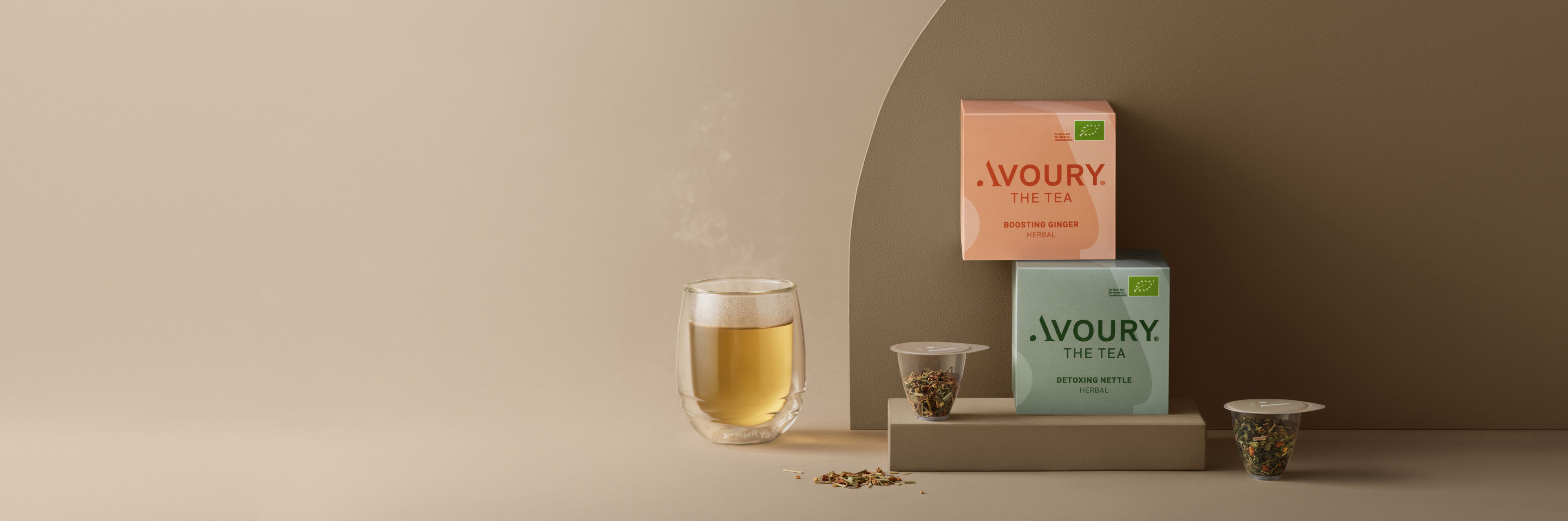 Two tea boxes labeled “Avoury The Tea,” along with two tea capsules and a steaming glas of tea. 