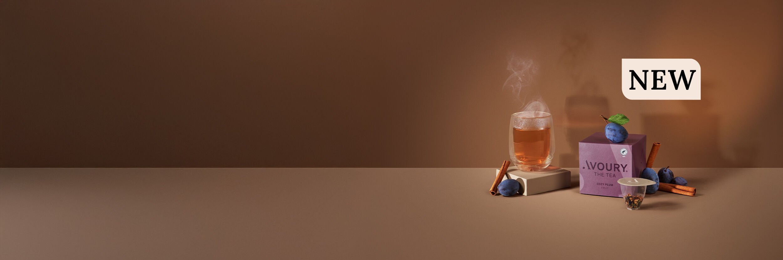 A steaming glass of tea, plums, cinnamon sticks, and a tea capsule arranged on a brown background.